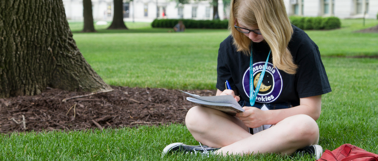 student sitting in grass writing in notebook